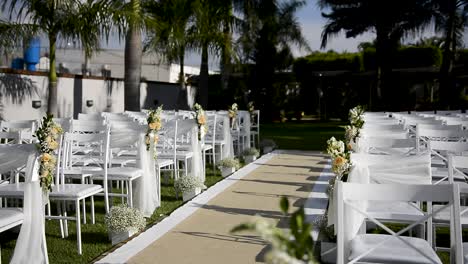 Outdoor-place-decorated-for-a-wedding-ceremony,-with-white-chairs-placed-over-green-grass,-carpet-in-the-center-aisle-decorated-with-flowers-and-veils,-view-from-the-altar
