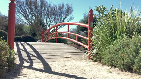Gravel-pathway-leading-to-red-bridge-over-pond,-Ju-Raku-En-Japanese-Garden,-Toowoomba,-Australia