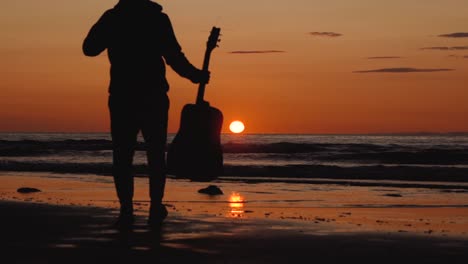 Man-running-with-guitar-in-back-sand-beach-at-sunset
