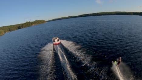 Slow-Motion---White-Man-Slalom-Water-Skis-from-White-Boat-on-Blue-Lake-in-Summer-Sunset-Light,-FPV-Drone-Aerial-Track-Follow