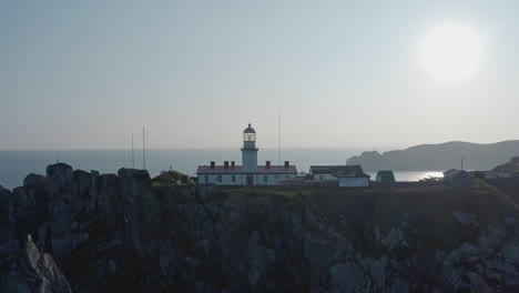 Slow-aerial-push-back-shot-of-Gamov-lighthouse-complex-standing-on-a-rocky-steep-cliff,-on-the-sunset