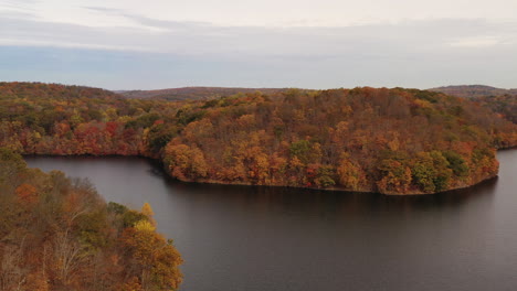 aerial-truck-shot,-left-over-the-reservoir,-as-the-drone-camera-focuses-on-the-orange-colored-tree-tops-of-autumn---mountains-on-a-cloudy-autumn-day-at-the-New-Croton-Dam-in-Westchester-County,-NY