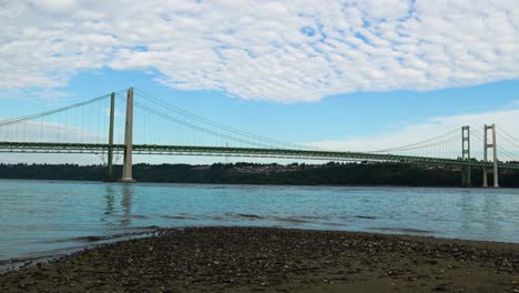 Time-lapse,-hyperlapse,-High-stratus-clouds-streaming-over-Puget-Sound-and-the-Tacoma-Narrows-Bridge,-Zoom-out-to-a-sand-and-pebble-beach