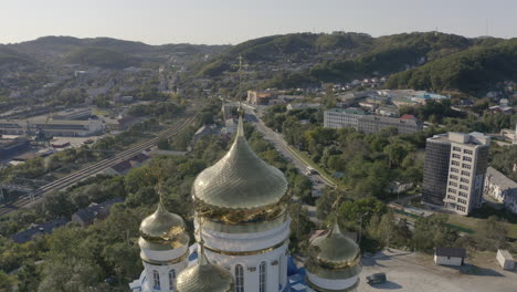 Slow-aerial-pivot-shot-of-orthodox-church-golden-domes-with-golden-crosses-on-top,-with-port-and-city-in-the-background-on-a-bright,-sunny-day