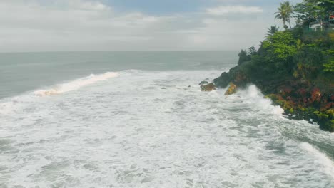 4k-Aerial-still-shot-of-Varkala-cliff-beach-with-lot-of-cafes,-moving-clouds-in-the-background-and-waves-crashing-at-the-bottom-of-the-cliff-in-the-morning