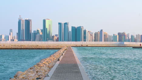 Soft-wind-blowing-blue-sea-waves-on-the-shore,-walkway-cement-and-rock-shore-with-modern-buildings-of-Manama,-Bahrain