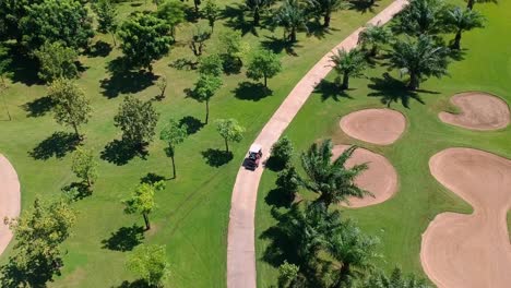 Golf-Cart-Driven-Through-Narrow-Road-Path-Inside-Huge-Golf-Course-Grounds---aerial-view