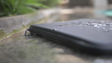 Close-up-of-a-black,-basic,-waterproof-laptop-case-with-lines-engraved-in-it-lies-on-the-side-of-a-pool-with-splashes-and-drops-of-water-on-it