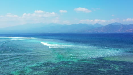 Beautiful-white-waves-foaming-over-turquoise-lagoon,-splashing-over-coral-reefs,-bright-blue-sky-with-white-clouds-in-Bali