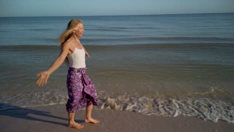 Looking-at-the-camera,-a-pretty-mature-woman-is-smiling-and-walking-on-the-beach-with-arms-outstretched-and-very-joyous