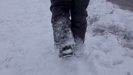 Young-child-walking-through-snow-at-side-of-road-wearing-grey-snowpants-and-boots-in-winter