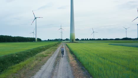 Young-female-is-riding-her-bicycle-on-a-dirt-road-through-a-green-field-of-windmills-renewable-energy-technology-park-with-tall-turbines-in-dutch-farmland-4k