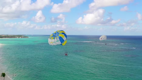 Scenic-view-of-two-people-parasailing-together-in-yellow-and-blue-parachute-by-sandy-Punta-Cana-resort-beach,-Dominican-Republic,-aerial