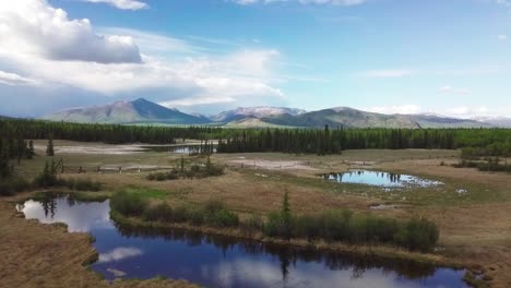 Spectacular-panoramic-nature-scene-of-outdoor-Yukon-landscape-and-flight-above-mirror-lake,-big-blue-expansive-sky-and-mountain-range-in-background,-Whitehorse,-Canada,-overhead-aerial-approach