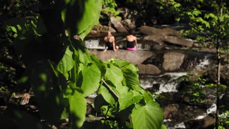 Two-woman-suntanning-on-the-rocks-in-the-middle-of-a-river-with-the-ruins-of-a-old-mill-in-the-background-in-Gatineau-Park,-Quebec