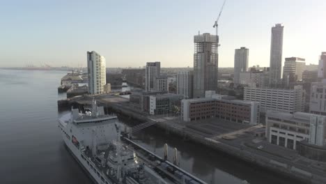 Liverpool-waterfront-aerial-view-royal-navy-military-ship-sunrise-high-rise-buildings-skyline-pull-back-descend