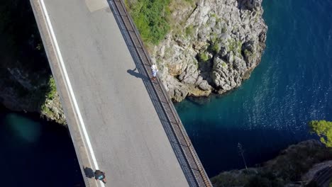 Fiordo-di-Furore-arch-bridge-with-a-man-with-hat-admiring-the-view-while-cars-pass-by,-Aerial-drone-top-orbit-shot