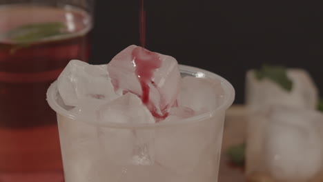 Studio-produc-shot-of-red-syrup-pouri-ng-in-to-plastic-cup-full-of-ice-with-lemonade-and-ice-in-background