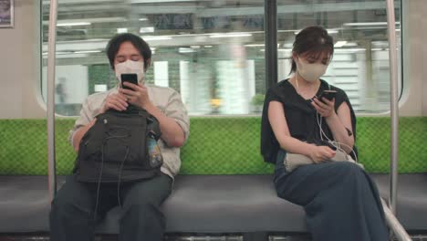 Hombre-Y-Mujer-Japoneses-Con-Máscaras-Y-Usando-Sus-Teléfonos-Inteligentes-Dentro-De-Un-Tren-En-Movimiento-En-Tokio,-Japón