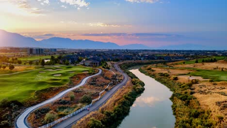 Aerial-hyperlapse-over-a-river-with-the-rising-sun-and-an-idyllic-community-with-a-golf-course,-commuter-train-and-a-city-in-the-valley