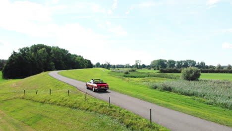 Ein-Rotes-Cabriolet-Auto,-Das-An-Einem-Sonnigen-Tag-Auf-Der-Straße-Durch-Die-Grüne-Landschaft-In-Den-Niederlanden-Fährt---Drohnenaufnahme