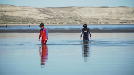 Two-kids-wearing-wetsuits-walking-on-the-shallow-waters-of-the-Patagonian-sea,-mountain-background---Wide-shot-UHD