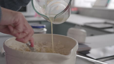 A-lady-making-dough-dough-in-the-kitchen,-she-pours-milk-into-the-mixture-in-a-ceramic-bowl-while-stirring