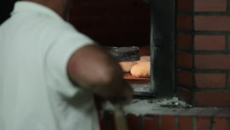 Male-Baker-With-Metal-Peel-Sliding-Dough-Of-Bread-Inside-The-Traditional-Brick-Oven-In-The-Bakery