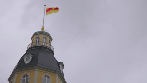 Die-Flagge-Eines-Turms-Vom-Schloss-Kalsruhe-An-Einem-Windigen-Und-Bewölkten-Tag,-Der-Eine-Traurige-Stimmung-Vermittelt