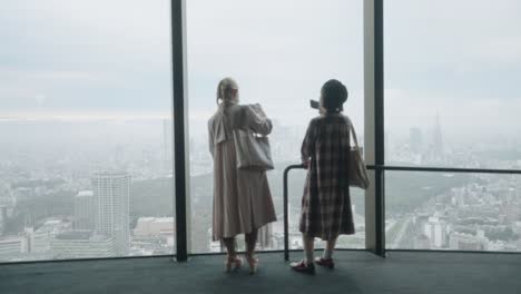 Zwei-Mädchen,-Die-Fotos-Der-Nebligen-Skyline-Von-Tokio-Durch-Die-Glaswand-Des-Shibuya-Himmels-Machen---Weitschuss