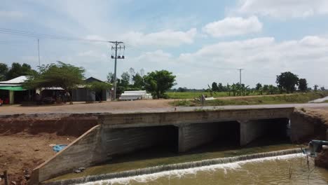 Dron-Volando-Lejos-Del-Puente-Sobre-El-Canal-De-Riego-En-El-Campo-De-Battambang,-Camboya