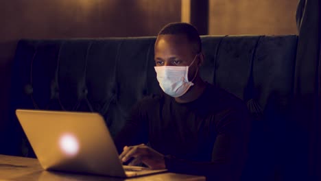 Male-working-on-a-laptop-in-a-dark-café-wearing-a-facemask