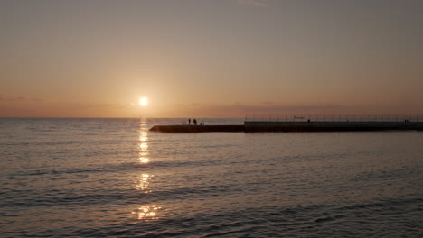 Zeitraffer-Des-Sonnenuntergangs-über-Dem-Meer-Mit-Wolkig-Orangefarbenem-Himmel-Und-Silhouette-Von-Menschen-Auf-Dem-Pier-Und-Booten-Am-Horizont