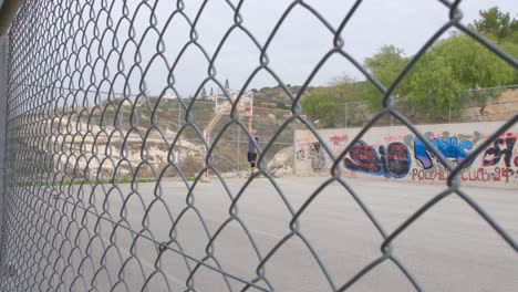 View-from-behind-a-chain-link-fence-of-a-man-playing-basketball-at-an-outdoor-court