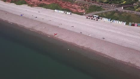 Flying-over-a-green-sea-along-a-pebble-beach-with-colorful-huts-and-boats,-AERIAL-STATIC-CROP