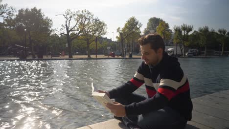 Nice-looking-young-man-sitting-by-a-canal-in-Amsterdam-concentrated-reading-a-book