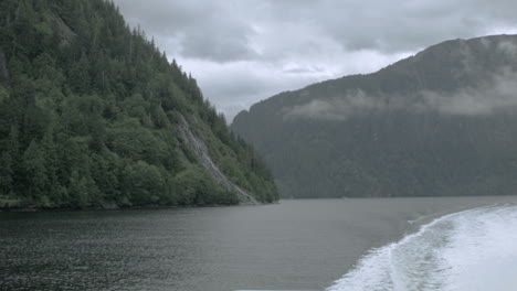 An-Alaskan-fjord-with-a-mountainside-in-the-foreground,-filmed-from-the-back-of-a-boat-creating-smooth-motion-with-a-settling-wake