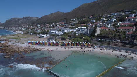 Drone-rises-to-reveals-beautiful-and-historically-coloured-beach-huts-of-St-James-beach-and-the-ocean-tide-pool-with-mountain-backdrop