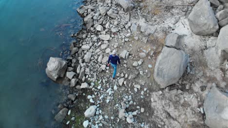 Aerial-shot-of-a-man-walking-along-rocky-lake-shore-in-mountains