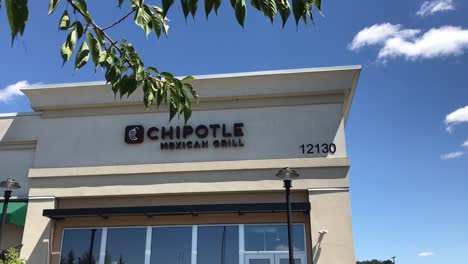 The-exterior-of-a-Chipotle-fast-casual-Tex-Mex-Restaurant-in-a-strip-mall-in-Portland,-Oregon,-USA-with-a-handheld-pan-down-through-some-trees
