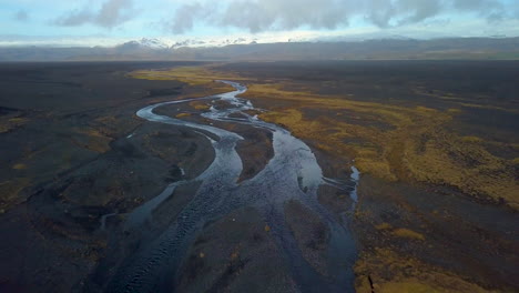 Drone-view-of-the-river-running-through-the-black-sand-beach-in-Iceland-with-mountains-in-the-background