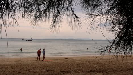 Schuss-Durch-Nadelbaum-Am-Strand-Von-Thailand-Mit-Glücklichen-Menschen,-Die-Auf-Dem-Sand-Spielen-Und-Tanzen