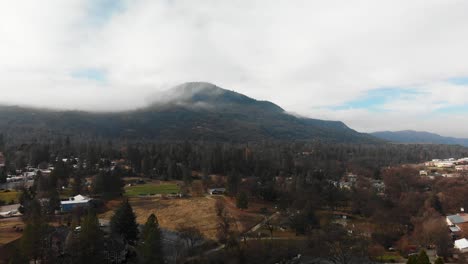 Aerial-descending-over-small-mountain-town,-cloud-covered-mountain-looming-in-background