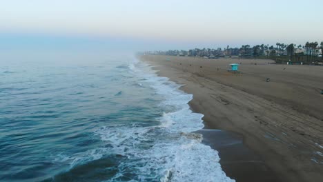 Ein-4K-Ariel-Blick,-Der-Sich-Bei-Sonnenaufgang-Die-Neblige-Küste-In-Huntington-Beach,-Kalifornien,-Surf-City,-USA-Hinunterbewegt,-Während-Surfer-Wellen-Fangen-Und-Familien-Ihre-Reise-Und-Ihren-Urlaub-Am-Strand-Genießen