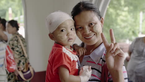 Glückliche-Birmanische-Mutter,-Die-Ihr-Verletztes-Kind-Hält,-Das-Ein-Friedenszeichen-Macht