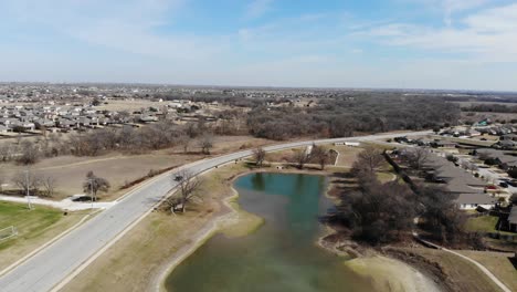 Pull-back-shot-of-drone-rising-above-a-neighborhood-pond-during-the-winter-with-a-brilliant-blue-sky-and-a-black-truck-going-by