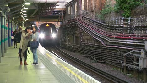 Tren-De-La-Línea-Del-Distrito-Subterráneo-De-Londres-Que-Llega-A-La-Estación-De-Whitechapel-En-El-Este-De-Londres,-Tower-Hamlets-Mientras-Dos-Mujeres-Esperan-En-La-Plataforma-En-Dirección-Oeste