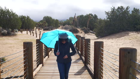 Pretty-woman-walking-across-a-bridge-with-a-blue-rain-umbrella-with-stormy,-cloudy-skies-above-SLOW-MOTION