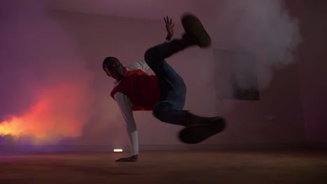 Slow-motion-shot-of-African-youth-in-red-shirt-doing-big-flares-with-his-leg-while-break-dancing-in-colorful-lights-and-smoke