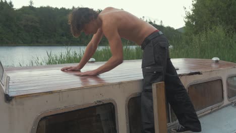 Oben-ohne-junger-Zimmermann,-Der-Die-Dachbeplankung-Eines-Holzbootes-Mit-Sandpapier-Von-Hand-Schleift